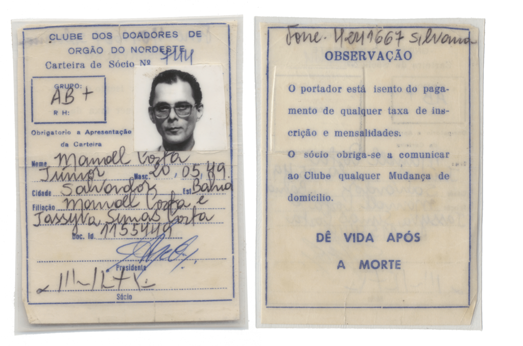 Documento do Clube dos Doadores de órgão do Nordeste. São duas folhas em formato
A1, antigas e amareladas, lado a lado, com letras escritas em azul.
À esquerda, a parte que representa a frente do documento, contém uma foto 3×4 no canto
superior direito, em preto e branco, de um homem de cabelo curto, sobrancelhas grossas e
rosto fino. Ele usa óculos de grau. Um texto posicionado mais à esquerda, no centro e na
parte inferior:
Clube dos doadores de órgão do Nordeste.
Carteira de Sócio n° 744
Grupo AB+
Obrigatório a Apresentação da Carteira
Nome: Manoel Costa Júnior
Nasc. 20/05/49
Cidade: Salvador Estado: Bahia
Filiação: Manoel Costa e Jassyra Simas Costa
Doc ID: 11554494
Mais abaixo: assinaturas do presidente e do sócio.
À direita, uma folha que representa o verso, escrito em caneta preta e letra cursiva Fone:
4241667. Silvana.
Abaixo, um texto datilografado em azul:
Observação. O portador está isento do pagamento de qualquer taxa de inscrição e
mensalidades: O sócio obriga-se a comunicar ao Clube qualquer mudança de domicílio. Dê
vida após a morte.