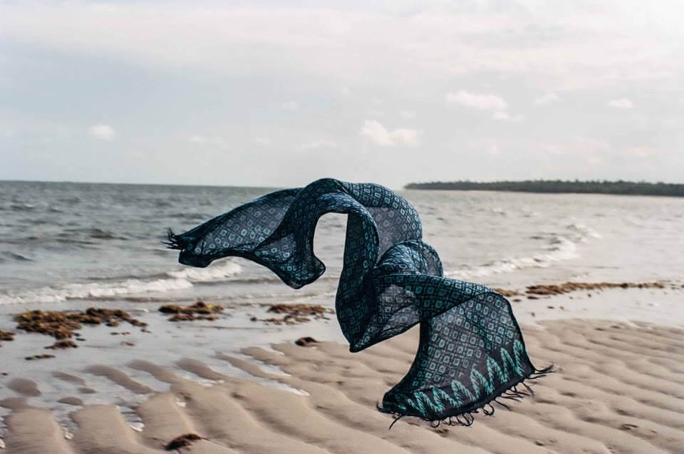 Fotografia paisagem de uma canga ao vento, semelhante ao voo de uma borboleta sobre a
faixa de areia da praia. A canga tem tons azuis, com delicadas estampas. No horizonte, o
mar com ondas brandas e a luz do sol amena, com o céu levemente nublado.