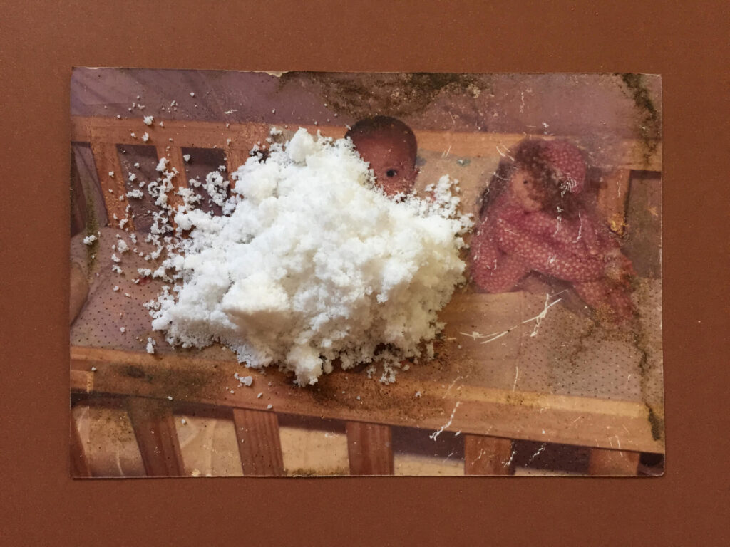 Fotografia retangular horizontal de um punhado de sal marinho granulado
sobre foto envelhecida, com manchas escuras de mofo e rasuras brancas. O
sal encobre a lateral direita do rosto e do corpo de um bebê de olhos castanhos, ao lado de uma boneca de pano sentada de perfil, ambos dentro de um berço de madeira. A foto está sobreposta a paspatur marrom. Roteiro de audiodescrição: Adamy. Consultoria: Ednilson Sacramento.
