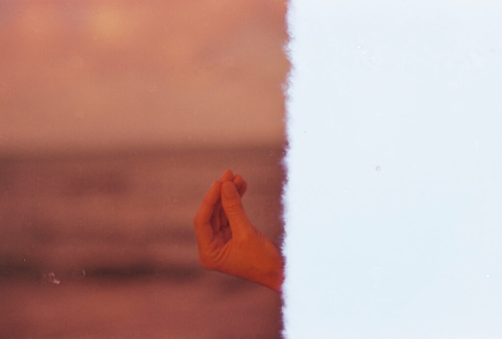 Papel fotográfico retangular dividido ao meio, na vertical. À esquerda, fotografia com a mão de uma pessoa de pele parda vista a partir do pulso, no canto
inferior direito. As pontas dos dedos estão unidas e tocam a ponta do polegar. O fundo em tons de marrom e âmbar está desfocado e o papel apresenta pequenas marcas de pingos de água e rasuras esbranquiçadas. O lado direito do papel é inteiramente branco. Roteiro de audiodescrição: Adamy. Consultoria: Ednilson Sacramento.
