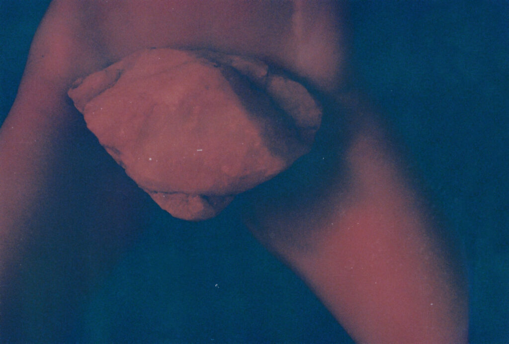 Fotografia retangular do quadril de uma pessoa de pele parda, magra, com as coxas entreabertas. Sobre a região genital, há uma pedra grande e volumosa, em formato triangular, com o vértice voltado para baixo. O fundo é azul escuro. Roteiro de audiodescrição: Adamy. Consultoria: Ednilson Sacramento.