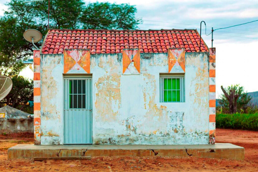 Foto paisagem. Em um chão de terra vermelha, a fachada de uma casa baixa com paredes brancas descascadas e telhas. No canto da casa, pilastras em laranja e branco, do lado esquerdo, uma porta de metal azul claro e uma antena pequena em formato de disco. Do lado direito, uma pequena janela quadrada azul com um fundo verde claro, nas telhas vermelhas, um metal com ponta curvada e um fio. No horizonte, uma vegetação baixa, árvores com poucas folhas, uma estrutura de concreto e céu azul com poucas nuvens. Roteiro de audiodescrição: Larissa Tavares. Consultora: Lara Souto.