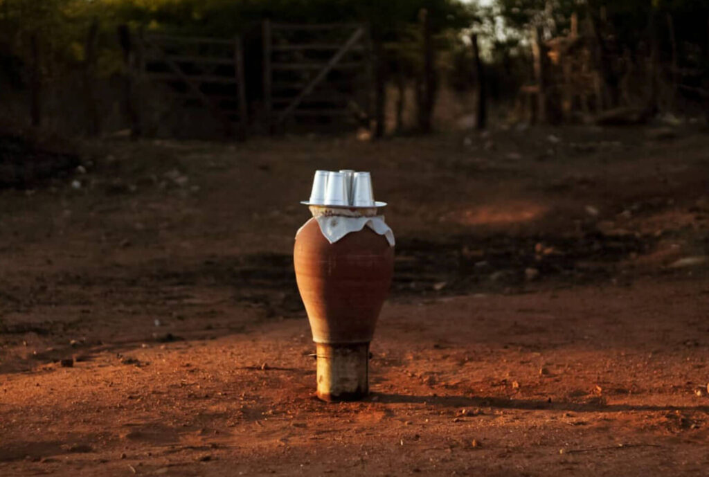 Foto paisagem. Em um chão de terra marrom, um vaso de barro sobre um balde enferrujado, em cima do vaso um pano branco e uma bandeja de 4 copos de alumínio. No fundo uma cerca torta e uma porteira levemente aberta. A luz do sol forma uma sombra do lado direito sobre a terra seca. Roteiro de audiodescrição: Larissa Tavares. Consultora: Lara Souto.