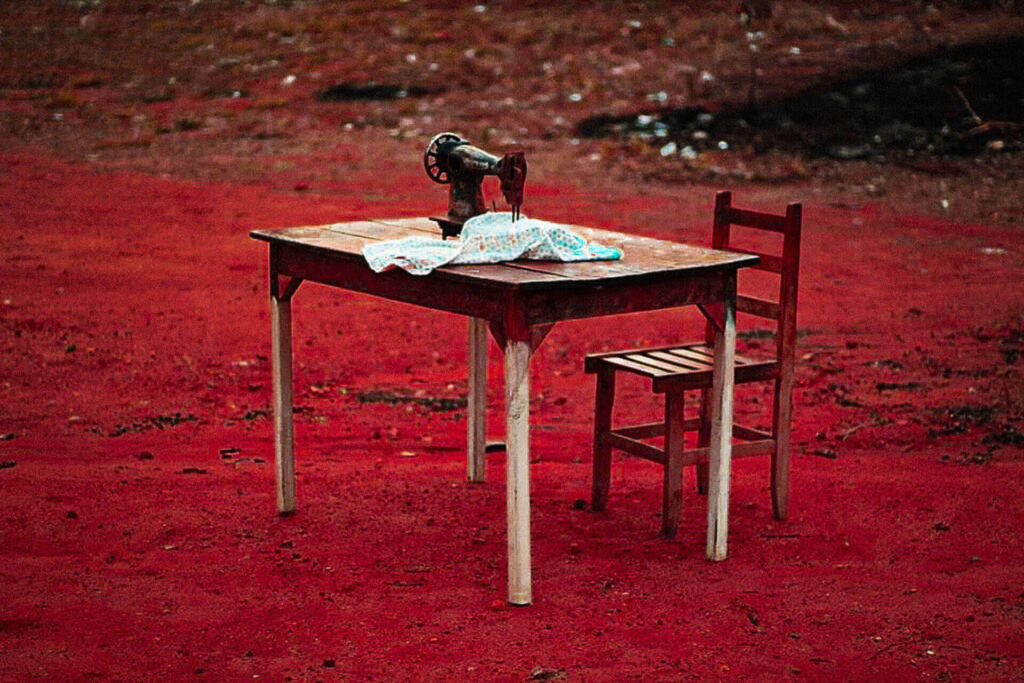 Foto paisagem. Em um chão arenoso vermelho, uma mesa retangular, com os pés esbranquiçados e uma cadeira de madeira escura. Sobre a mesa, um tecido de chita com estampa em azul, uma máquina de costura antiga preta. À direita, uma rampa pedregosa. Roteiro de audiodescrição: Larissa Tavares. Consultora: Lara Souto.