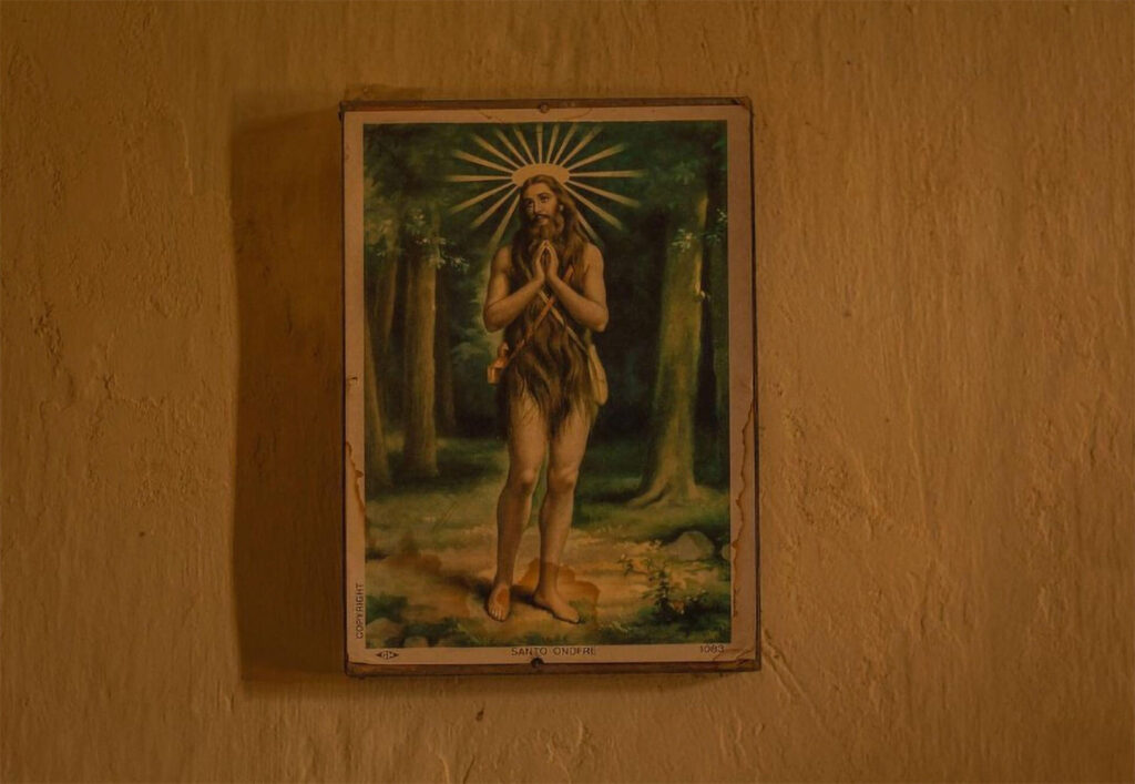 Foto retrato. Em tom amarelado de um quadro colorido de Santo Onofre em uma parede com marcas de reboco. Na obra, o santo está de pé em uma floresta. É um homem de pele clara, cabelos e barba longos, usa uma coroa dourada. Os cabelos de Onofre cobrem o corpo são ondulados e vão até os flancos. Roteiro de audiodescrição: Larissa Tavares. Consultora: Lara Souto.