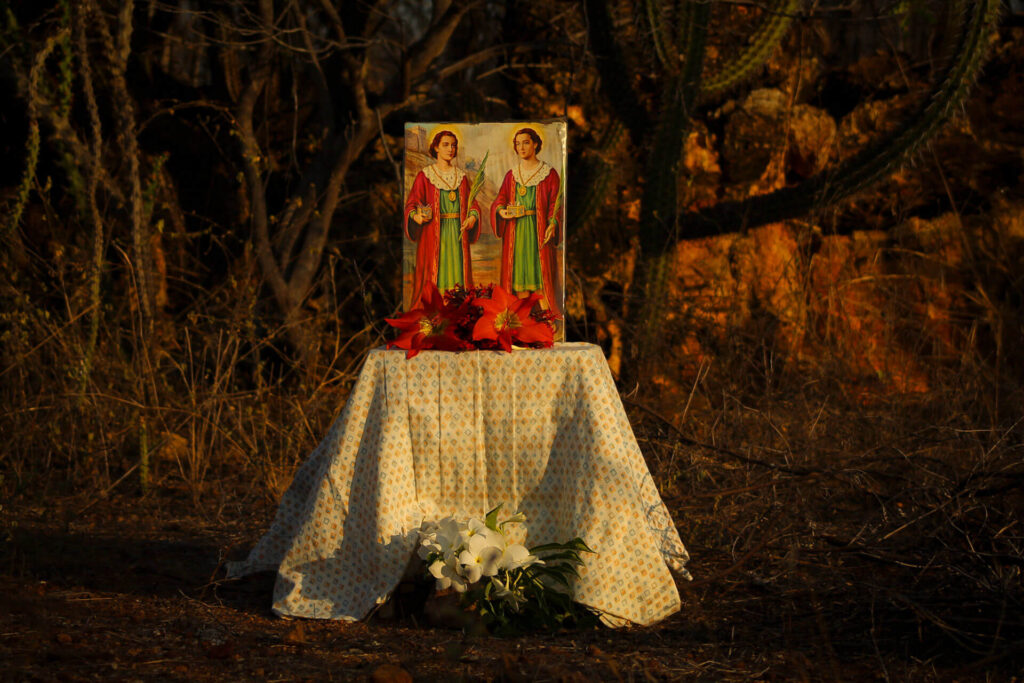 Foto paisagem. Em uma mesinha coberta por uma toalha branca com estampa de losangos em amarelo e azul, o quadro colorido de corpo inteiro de São Cosme e Damião. No quadro, os santos gêmeos estão de pé, seguram um ramo de palmeira, uma caixinha dourada com cruz vermelha; eles são brancos e tem o cabelo castanho claro penteados para trás. Os irmãos usam uma túnica verde clara com detalhes em dourado e um kimono vermelho longo. Na mesa à frente do quadro flores vermelhas, na base da mesa no chão de terra flores brancas. Ao redor, uma vegetação seca com galhos e cactos. Roteiro de audiodescrição: Larissa Tavares. Consultora: Lara Souto.