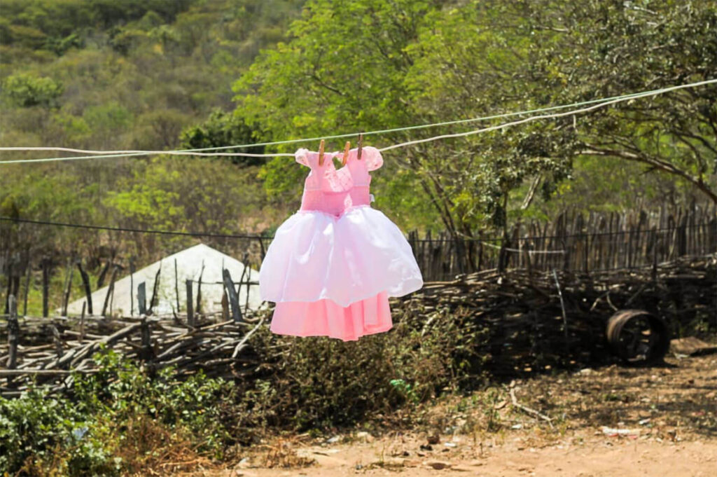 Fotografia em paisagem. Um vestido de criança rosa claro com saiote branco está pendurado em um varal. Ao fundo uma cerca de galhos finos e alguns gravetos amontoados no chão de terra. A luz do sol ilumina as folhas verdes e revela uma textura lisa da densa vegetação do espaço. Do lado esquerdo, há um telhado triangular de cor cinza claro. Roteiro de audiodescrição: Larissa Tavares. Consultora: Lara Souto.