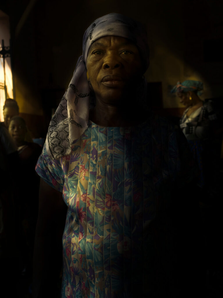 Foto retrato de uma senhora preta. Ela usa um lenço lilás com aspecto de cetim e está de vestido reto branco com estampa floral em lilás, azul e rosa. A senhora está em um contorno preto sombreado, do lado esquerdo um menino e uma mulher preta e do lado direito uma mulher mais velha preta; ela usa um lenço azul com um laço na ponta. Do lado direito uma luz natural entra no espaço fechado. 