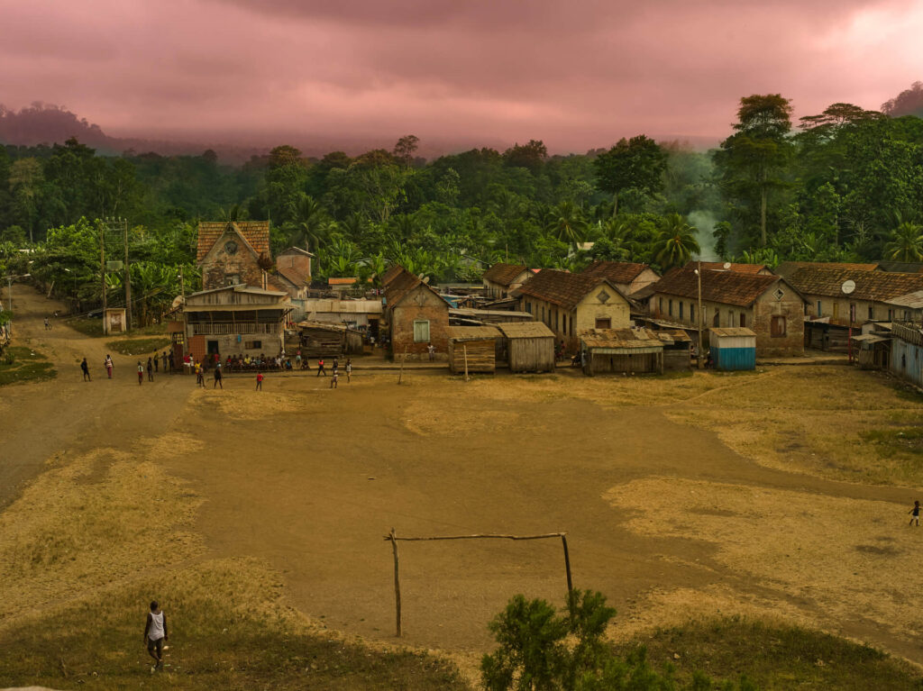 Foto paisagem aérea de uma campo de terra. O espaço tem   traves de madeira, um grupo de pessoas do lado esquerdo em uma das traves e uma pessoa preta de regata branca na outra. Ao fundo, casas baixas com telha vermelha, atrás das casas uma vegetação densa. O céu está lilás com nuvens e neblina. Uma fumaça branca sai de uma das casas do lado direito do campinho. 