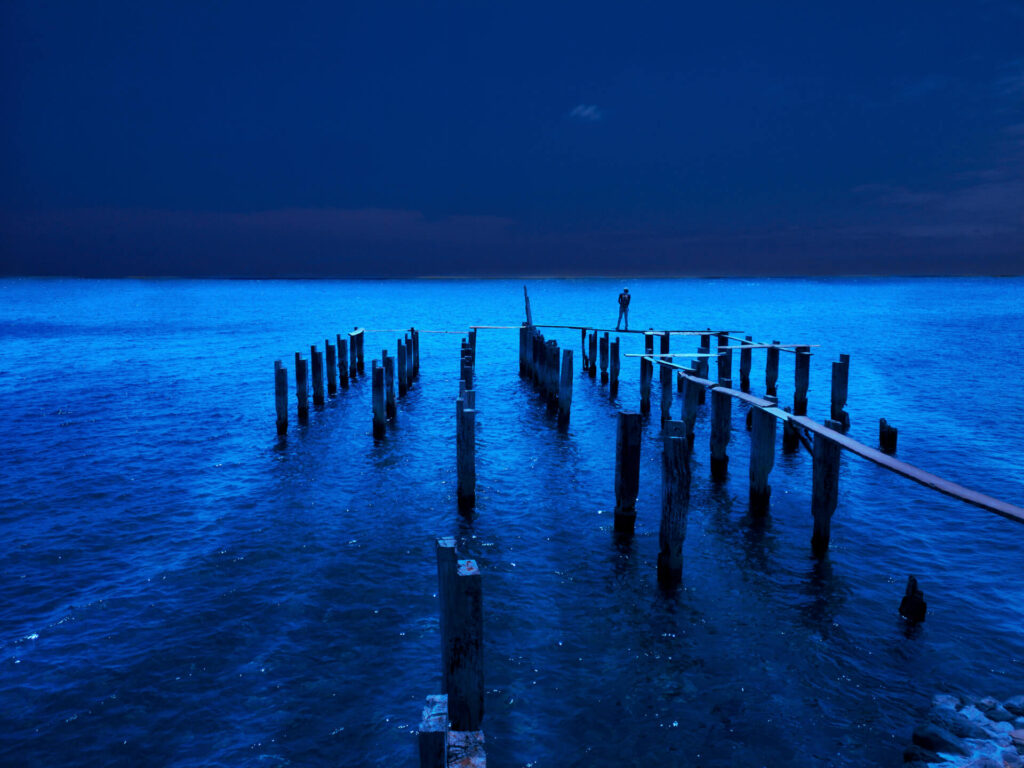 Foto paisagem de um pier em um mar azul escuro. O pier é de madeira estreita com diversas estacas organizadas em bloco, uma pessoa está parada no limite do pier. O céu é azul escuro sem nuvens. 
