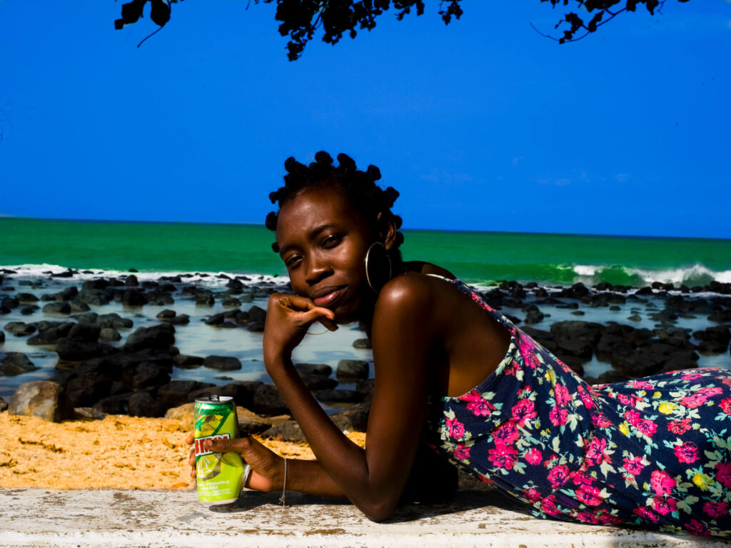 Foto paisagem de uma mulher posando para a foto em uma praia. Ela tem os cabelos em um penteado de coquinhos bantum, sobrancelhas arqueadas, olhos levemente fechados, nariz núbio, lábios grossos e rosados na parte inferior. A jovem é magra e está deitada de barriga para baixo em uma encosta com aspecto de concreto, é vista da cintura pra cima, usa um vestido preto com estampa floral em rosa, está com a mão direita no queixo e a mão esquerda amassando uma latinha verde. Ao fundo um céu azul, um mar verde claro, a espuma da água se encontra e diversas pedras espalhadas e a areia bege que se encontra como concreto onde está a jovem. O sol ilumina a pele negra e o sorriso tímido da mulher. 