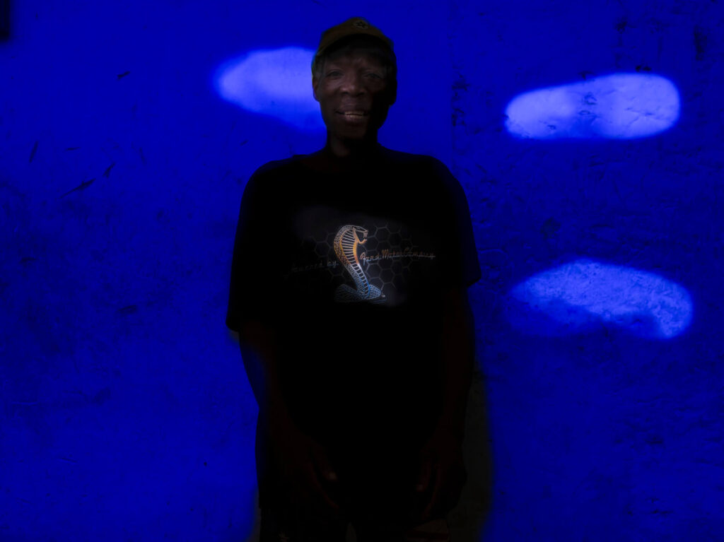 Foto retrato de um homem negro posando com o braço direito para trás e o esquerdo ao lado do corpo, visto da cintura pra cima, o cabelo é grisalho nas laterais, usa boné marrom; tem as sobrancelhas finas, olhos pequenos, lábios grossos e um leve sorriso no rosto.  Ele usa uma camiseta preta com um desenho de uma cobra cascavel e calça preta. Ao fundo uma parede com aspecto de concreto e iluminação azul. 