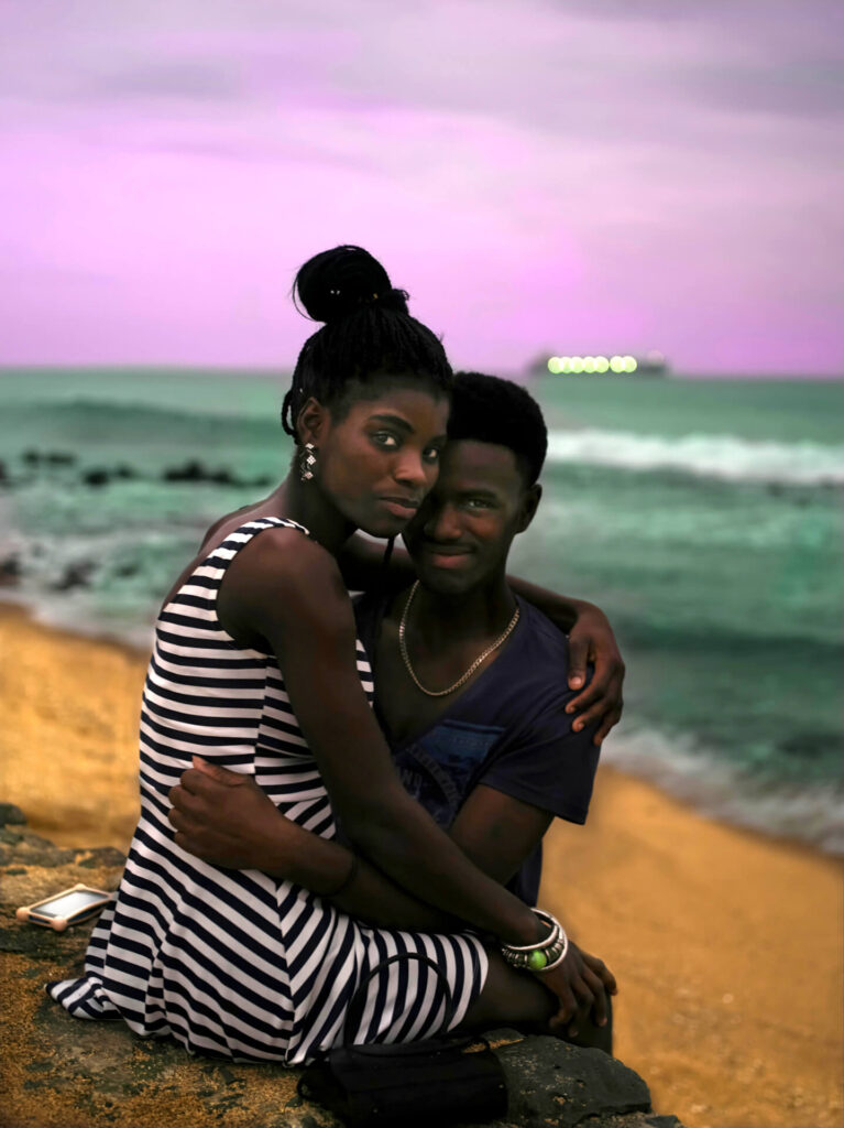 Foto retrato de um homem e uma mulher negros abraços em uma praia. Ela é uma mulher preta, com cabelos trançados e um coque alto, com sobrancelhas grossas, olhos pretos e nariz pequeno, lábios médios; ele tem os cabelos crespos curtos, sobrancelhas finas arqueadas, olhos pequenos, nariz núbio e lábios grossos. A mulher está sentada em um muro de pedras na beira da praia, o homem está abraçado entre as pernas da moça, ambos posam para a foto. Ela usa um vestido curto com linhas brancas e pretas, brincos e argolas com bolinhas; ele veste uma camiseta decotada azul escuro e um colar dourado. No muro um celular com a tela virada para cima, ao fundo um céu azul, com nuvens com fundo lilás, a água do mar tem cor esverdeada e uma onda quebra na beira da área amarela. 