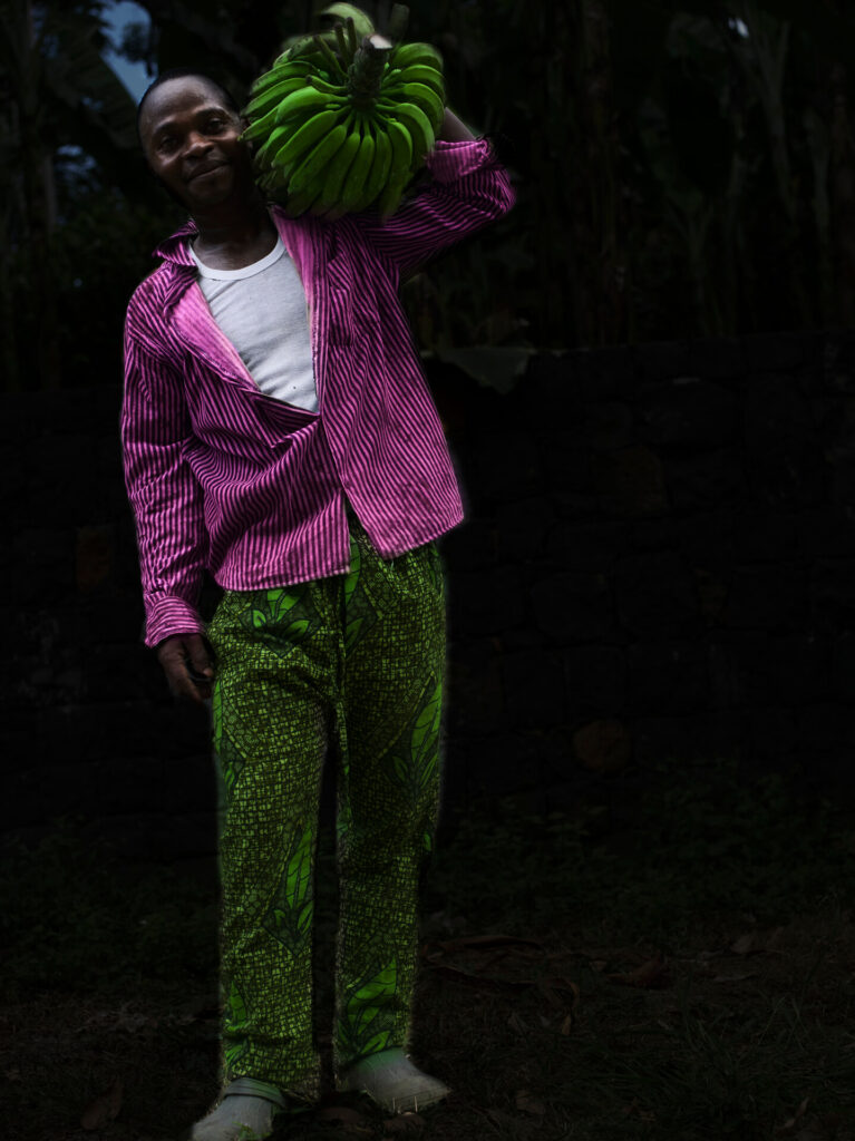 Foto retrato de homem preto de corpo inteiro segurando um cacho de banana verde. O homem está em uma mata aberta, há uma sombra que parece noite; ao fundo uma vegetação densa e no solo folhas secas. A imagem do homem de pé está contornada por uma luz fina esbranquiçada. Ele tem sobrancelhas finas, olhos grandes e pretos, nariz núbio e lábios grossos. Ele carrega o cacho no ombro direito. Veste uma blusa branca, uma camisa rosa com linhas pretas, calça verde claro com estampa de folhagens e meias cinzas. 