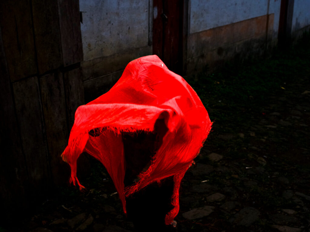 Foto paisagem com luz baixa. Um objeto está coberto por um tecido de morim vermelho. O objeto está em uma rua de pedras, ao fundo uma parede branca suja e duas portas de madeira com manchas escuras. 