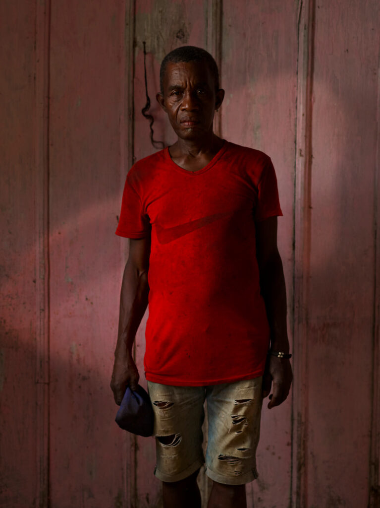 Foto retrato de um homem preto visto dos joelhos para cima. Ele tem os cabelos levemente raspados com entradas, tem marcas de expressão na testa, sobrancelhas espessas, nariz núbio e lábios grossos; usa uma camiseta vermelha surrada e uma bermuda jeans rasgada e suja; o homem segura um tecido azul na mão direita e uma pulseira dourada. Ao fundo uma parede de madeira rosa clara. 