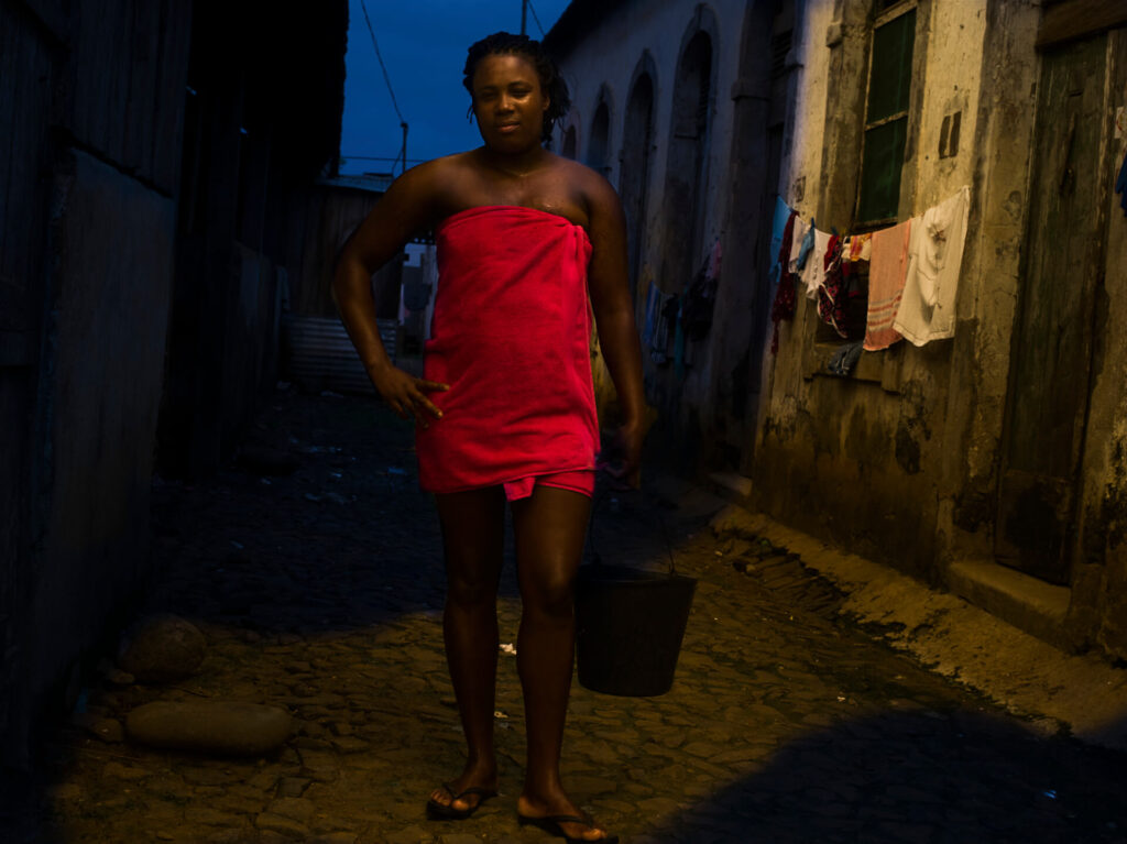 Foto retrato de uma mulher preta de corpo inteiro. Ela está em uma rua estreita de pedras (viela com asfalto de pedras), é preta de cabelos trançados e amarradores, tem sobrancelhas arqueadas, olhos pequenos e lábios grossos. A mulher está com a mão direita na cintura e segura um balde com a outra. Ela está enrolada em uma toalha de banho rosa e usa chinelos pretos. Ao fundo um céu azul escuro, dos lados muros brancos com manchas escuras; na parede da direita, panos e roupas em um varal. 