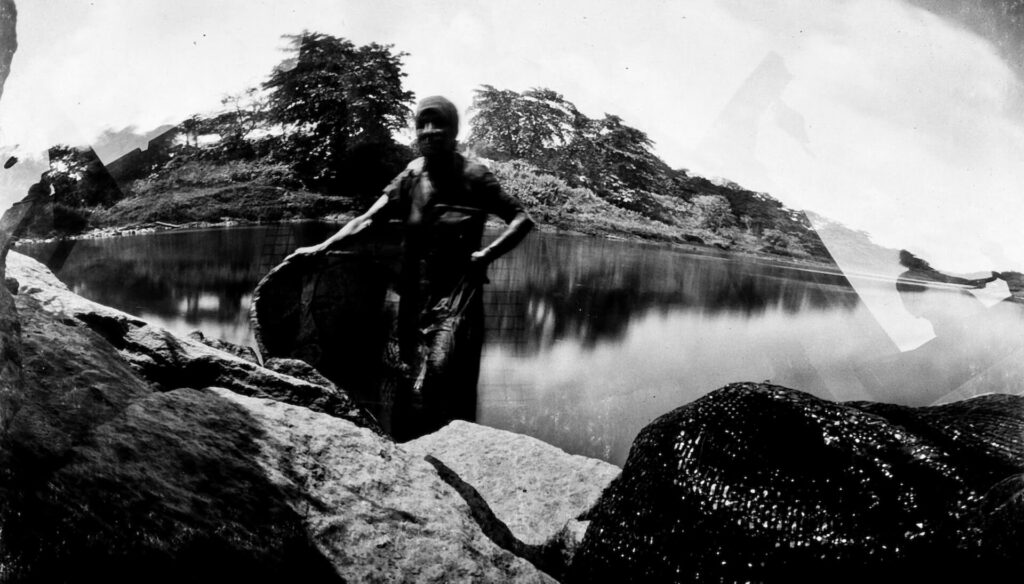 Fotografia paisagem, com sobreposição de fotos.  Entre o rio e as rochas da margem,  uma pessoa, com aparência feminina. Ela é magra e usa algo na cabeça, assemelha-se a um lenço e segura uma espécie de tecido.  No canto direito da rocha, há outro tecido preto, com pontos iluminados.  Ao longe, árvores e vegetação costeira. 