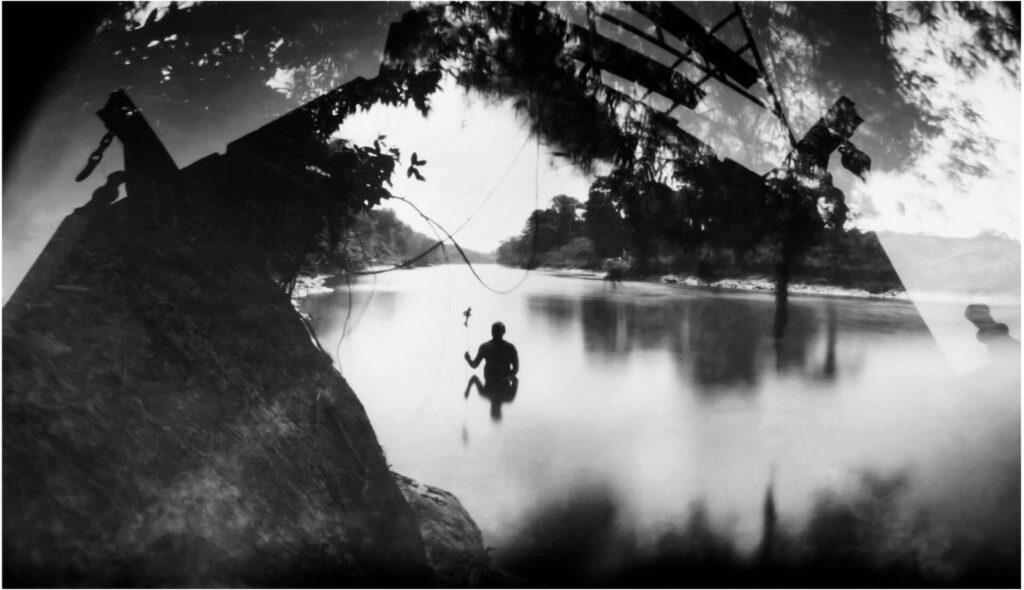Fotografia paisagem, com sobreposição de fotos. No centro, uma pessoa no rio, vista da cintura para cima, com a imagem refletida na água. Ela segura com a mão direita algo semelhante a uma vara de pesca. A pessoa tem o cabelo curto e corpo robusto. O rio é amplo, com vegetação costeira. No entorno da fotografia, perpassadas na parte superior, projeções de sombras de  pedra,  de copa de árvores e de estrutura de madeira. As folhas da árvore também estão refletidas nas águas do rio. 