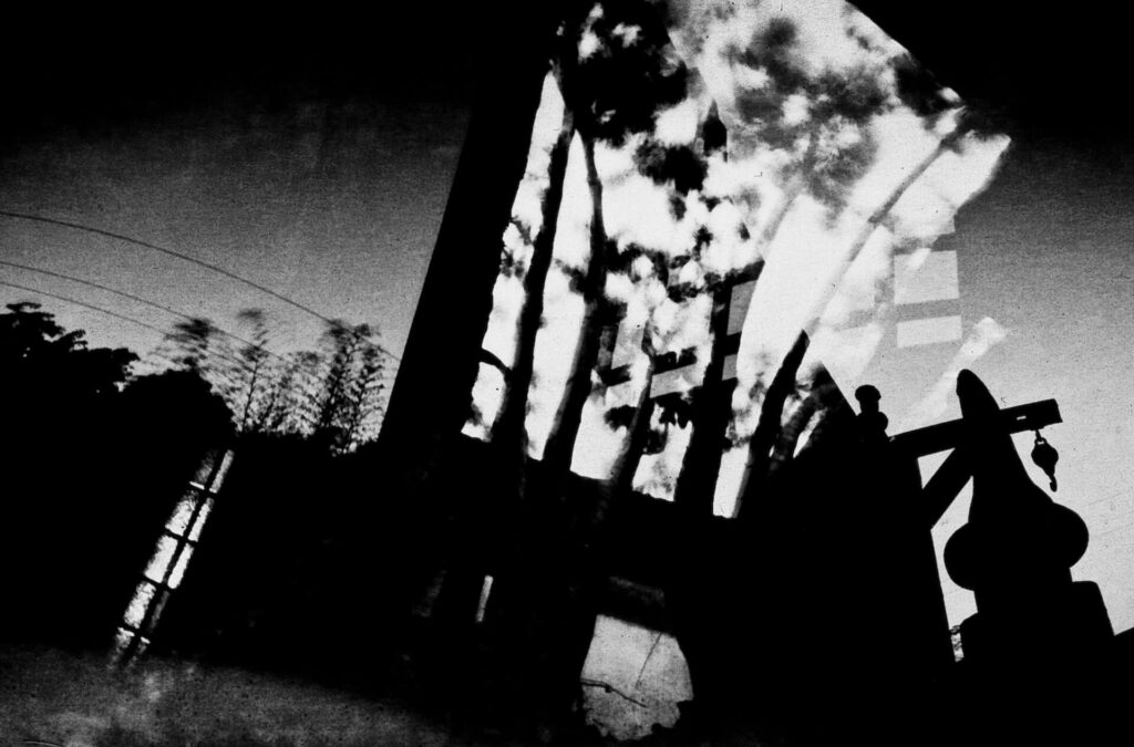 Fotografia paisagem, com sobreposição de fotos. A imagem se assemelha a uma rua com construções e sombras de árvores projetadas na parte superior.  Ao longe, há fios elétricos em arco e copas de árvores. 