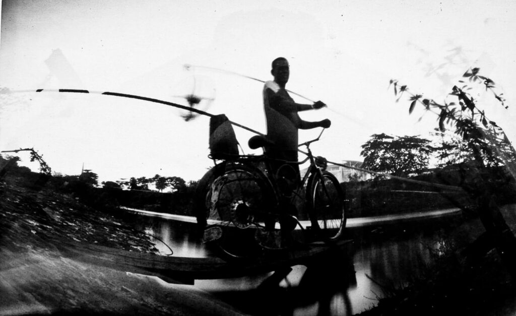 Fotografia paisagem. No centro, uma pessoa de perfil ao lado de uma bicicleta. Com a mão direita ela segura o guidão e com a esquerda uma vara de pesca. A pessoa tem cabelo curto, aparência  masculina e usa camisa. Ao redor, vegetação rasteira. 