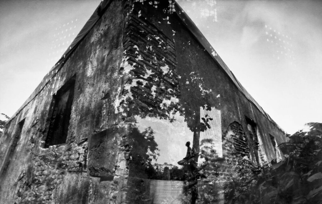 Fotografia paisagem. Detalhes da quina de uma alta fachada  em ruínas, com paredes sem reboco. No centro da quina, sobrepõe a imagem de uma pessoa de perfil, de cabelo curto, corpo magro, assemelha-se a um rapaz,  encostado no tronco de uma árvore, com a cabeça levemente erguida e um dos joelhos flexionado. Acima, projeção das sombras da copa da árvore. 