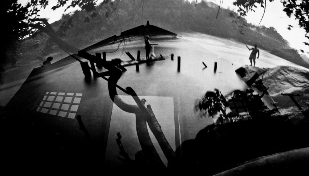 Fotografia paisagem, com sobreposição de fotos.  Nas águas do rio, reflexos de uma casa e um tronco de árvore. A casa tem  janela quadriculada e portal quadrado e o tronco está na transversal sobre a casa. Ao longe,  no centro, uma pessoa sobre a canoa, com uma rede de pesca e nas laterais, de um lado o rosto humano e do outro lado, alguém com aparência jovem e forte, com uma vara de pesca posicionada para o rio. Ao redor, pedras, vegetação e árvores. 
