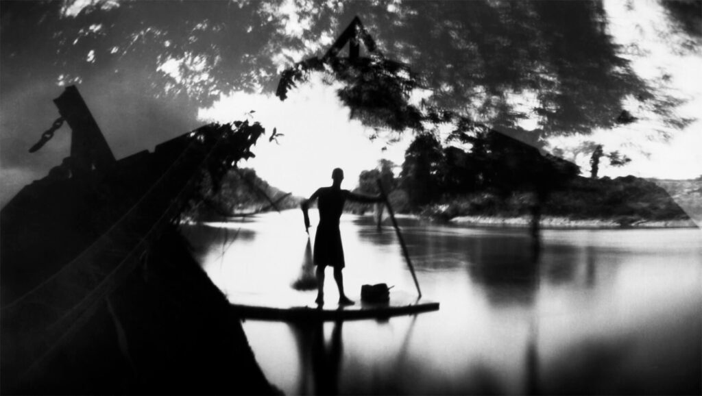 Fotografia paisagem, com sobreposição de fotos. No centro, o rio e uma pessoa sobre uma canoa. Ela é alta, forte e de cabelo curto, que segura uma rede de pesca e um remo de pau. Na parte superior, há reflexos de copas  de árvores e no entorno, fragmentos de construção. Ao longe, vegetação costeira. 
