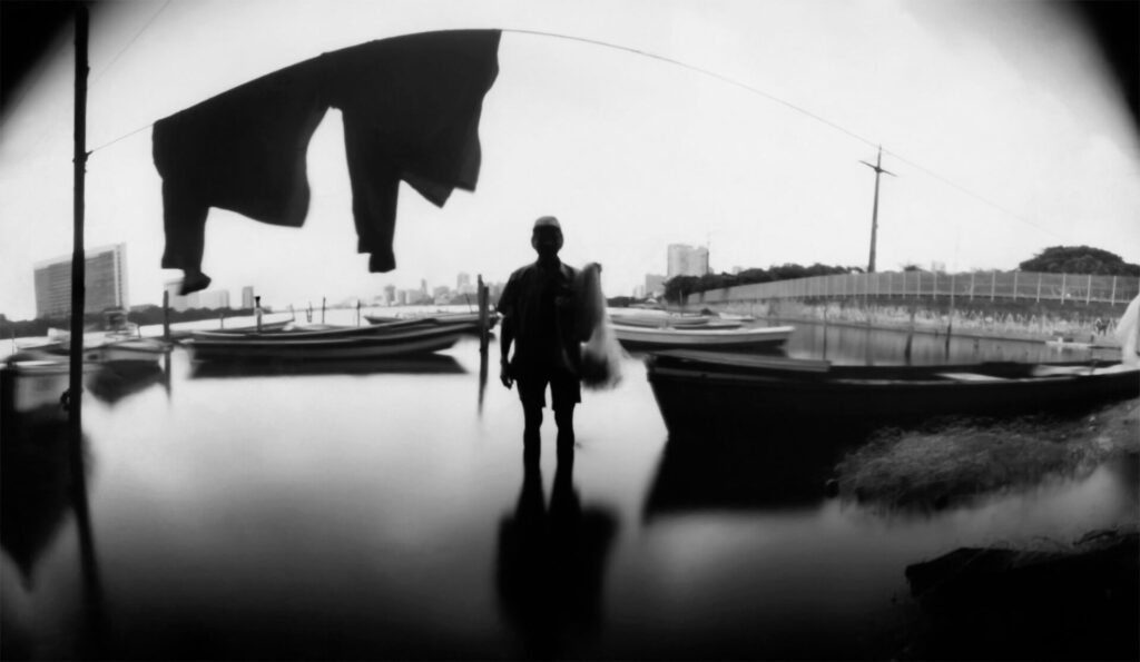 Fotografia paisagem. No centro, uma pessoa, com aparência masculina , na margem do rio. Ela aparenta ser um senhor de meia-idade, segura uma rede de pesca.  Na parte superior, há um varal com roupas penduradas. E ao redor, alguns barcos e à direita uma ponte. No horizonte, várias construções, uma delas se assemelha à arquitetura do prédio da Prefeitura da Cidade do Recife. 