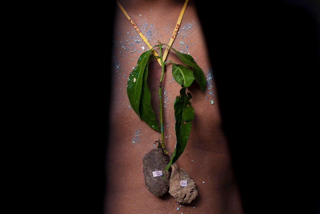 Fotografia retrato. Sobre fundo preto, close do tórax e abdômen de pele preta, com pontos de brilho azul e branco espalhados pelo corpo. Sobre o tórax, um colar de corda amarela, com pingente de folhas verdes e de raízes marrom e bege. Nas laterais da corda amarela, escrito em letras vermelhas: ainda pulsa. A musculatura do abdômen é levemente definida. Roteiro de audiodescrição: Vanessa Marques. Consultoria: Jéssica Morais.