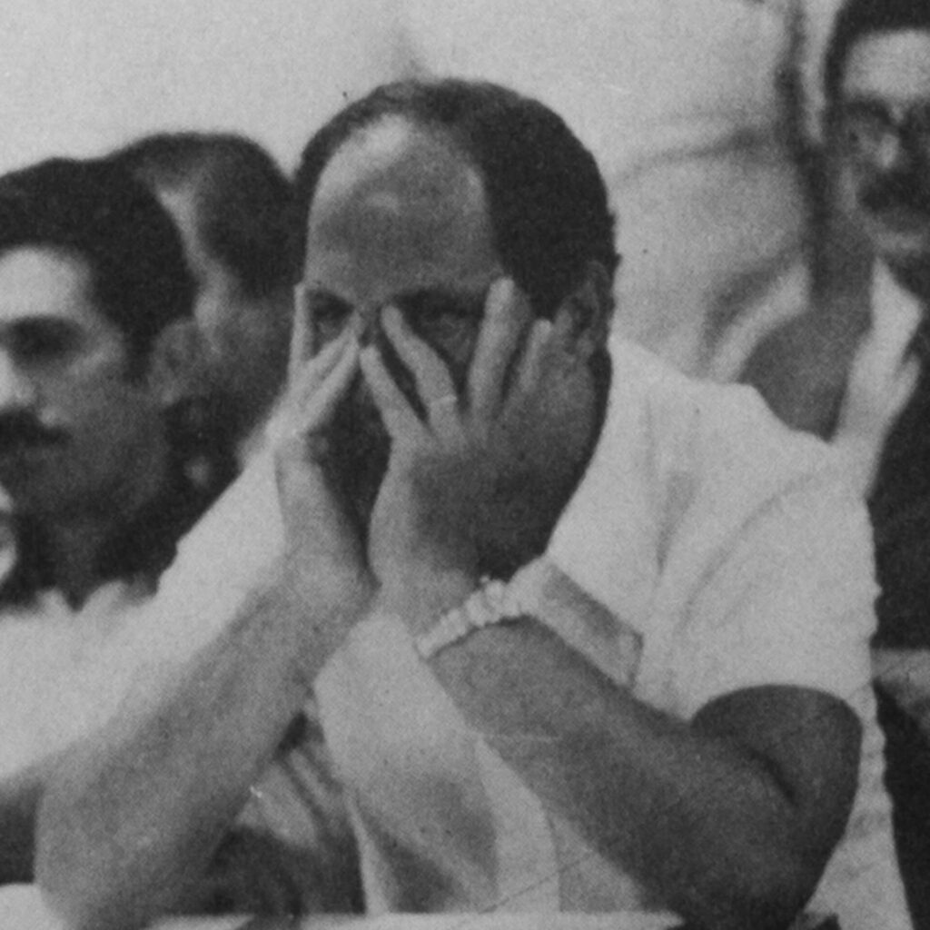 Foto de uma fotografia antiga, preta e branca, na vertical de um homem sentado
atrás de uma mesa com os cotovelos apoiados na mesa e as mãos no rosto que
deixa seus olhos pretos à mostra. Ele tem a testa avantajada, cabelos curtos, usa
uma camisa de botões de mangas curtas e uma aliança no dedo anelar da mão
esquerda e um relógio no punho esquerdo. Do lado esquerdo dele, dois homens
sentados e ao fundo outro homem sentado.