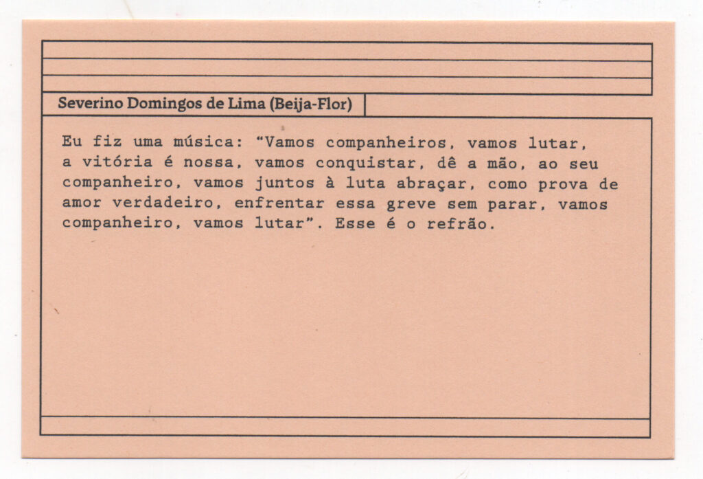 Fotografia de uma ficha horizontal cor de rosa claro com cinco pautas pretas na
parte superior. Abaixo delas, em letras pretas, o depoimento de Severino Domingos
de Lima (Beija-Flor): “Eu fiz uma música: ‘Vamos companheiros, vamos lutar, a
vitória é nossa, vamos conquistar, dê a mão ao seu companheiro, vamos juntos à
luta abraçar, como prova de amor verdadeiro, enfrentar essa greve sem parar,
vamos companheiro, vamos lutar’. Esse é o refrão.” No rodapé, duas pautas pretas
horizontais.
