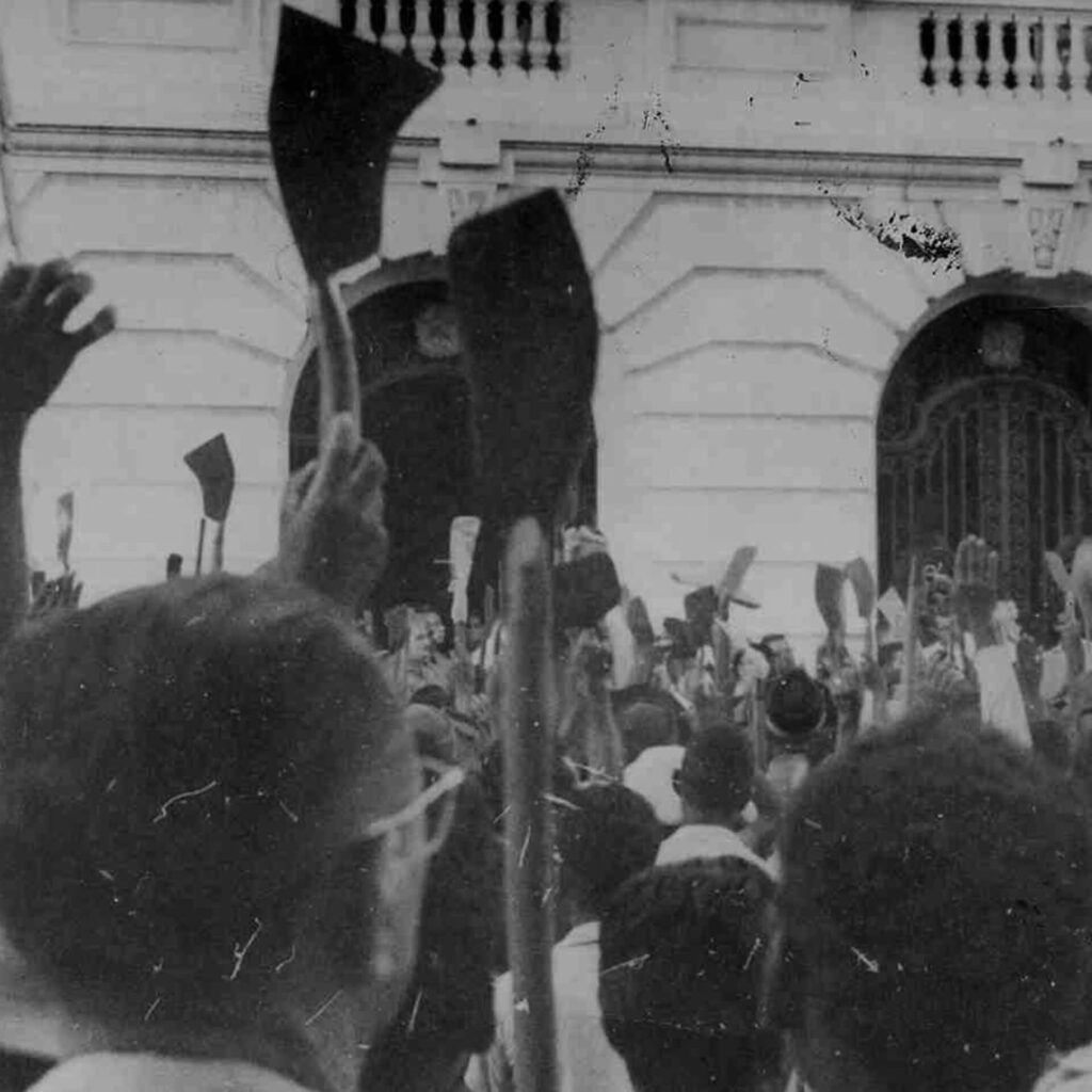 Foto de uma fotografia antiga, preta e branca, na vertical de trabalhadores de
costas, eles levantam umas foices na frente da fachada de um casarão com dois
portões largos de ferro.