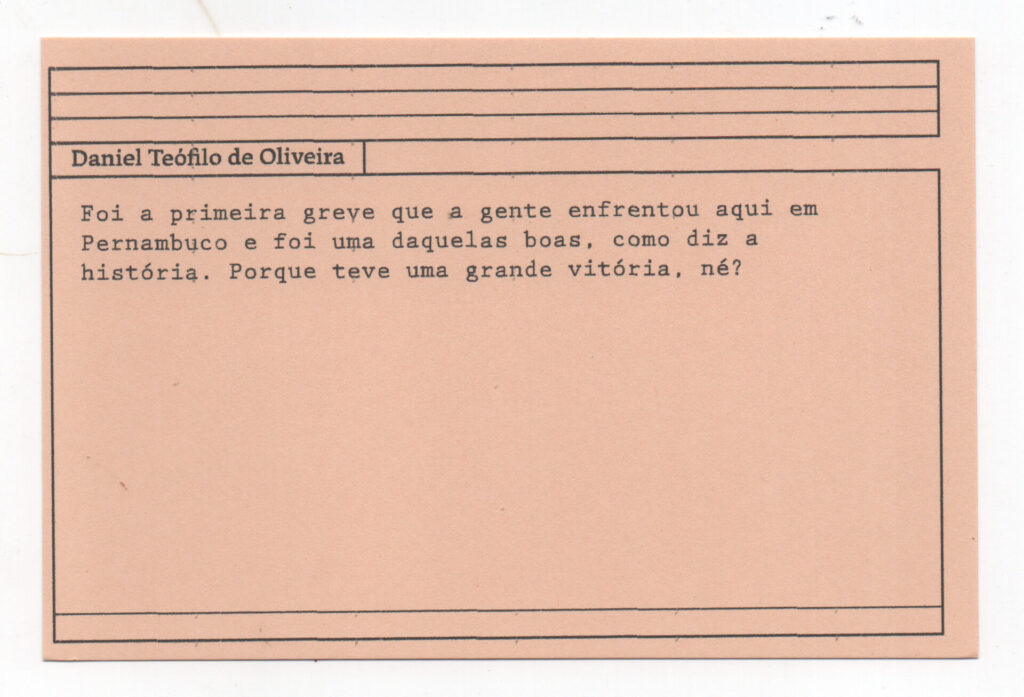 Fotografia de uma ficha horizontal cor de rosa claro com cinco pautas pretas na
parte superior. Abaixo delas, em letras pretas, o depoimento de Daniel Teófilo de
Oliveira, em letras pretas: “Foi a primeira greve que a gente enfrentou aqui em
Pernambuco e foi uma daquelas boas, como diz a história. Porque teve uma grande
vitória, né?” No rodapé, duas pautas pretas horizontais.