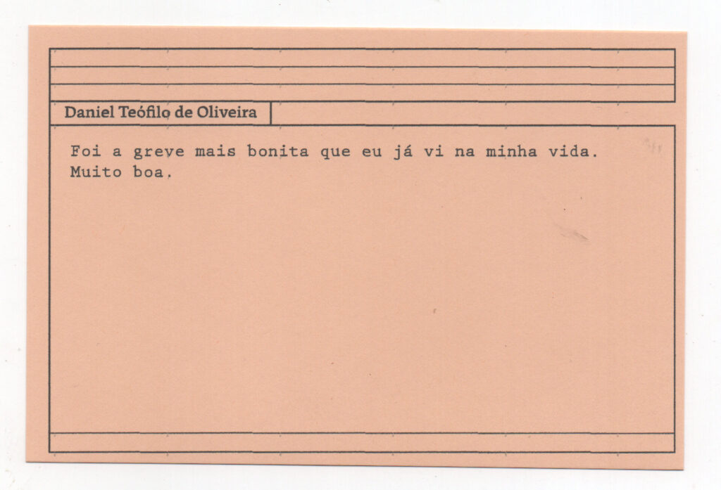 Fotografia de uma ficha horizontal cor de rosa claro com cinco pautas pretas na
parte superior. Abaixo delas, em letras pretas, o depoimento de Daniel Teófilo de
Oliveira, em letras pretas: “Foi a greve mais bonita que eu já vi na minha vida. Muito
boa.” No rodapé, duas pautas pretas horizontais.