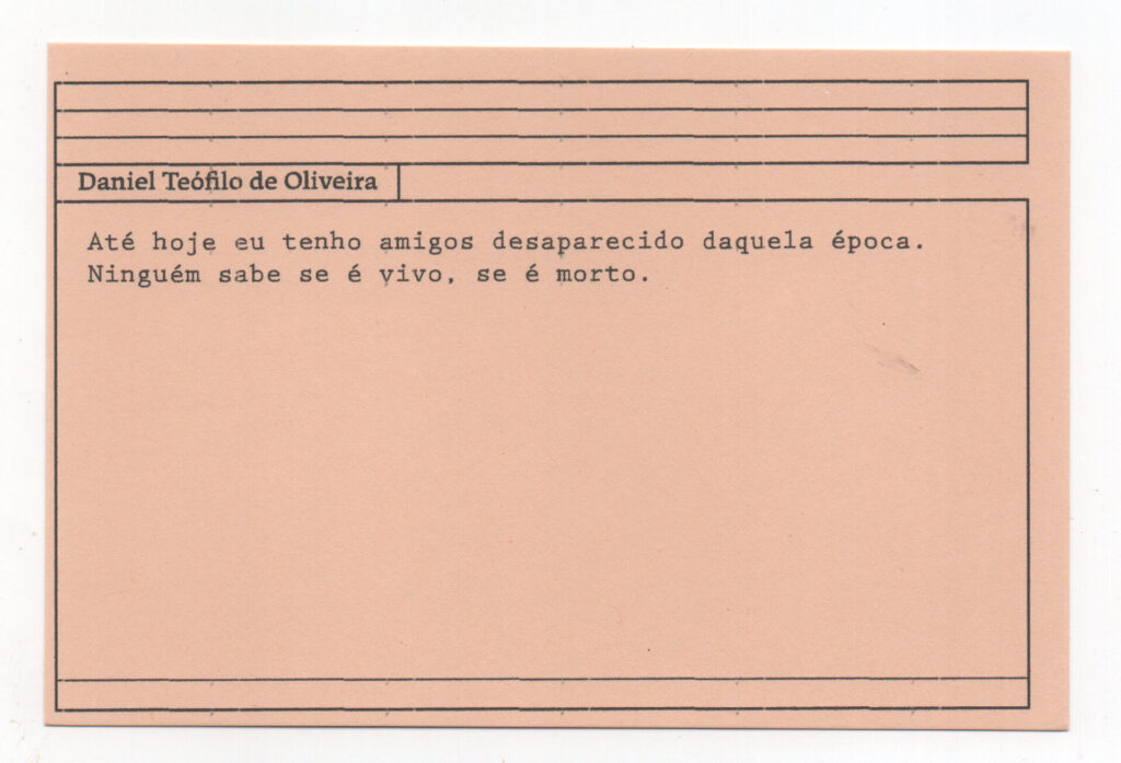 Fotografia de uma ficha horizontal cor de rosa claro com cinco pautas pretas na
parte superior. Abaixo delas, em letras pretas, o depoimento de Daniel Teófilo de
Oliveira, em letras pretas: “Até hoje eu tenho amigos desaparecido daquela época.
Ninguém sabe se é vivo, se é morto.” No rodapé, duas pautas pretas horizontais.