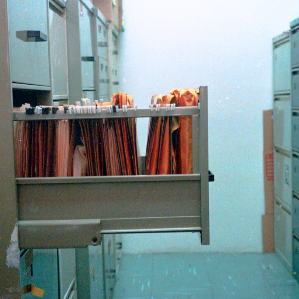 Fotografia na horizontal de um corredor de armários de aço na lateral esquerda e
direita. O armário, do lado esquerdo, tem a segunda gaveta cinza aberta com umas
pastas arquivo suspensas e uns envelopes pardos. À direita, armários com gavetas
fechadas. Ao fundo uma parede branca.