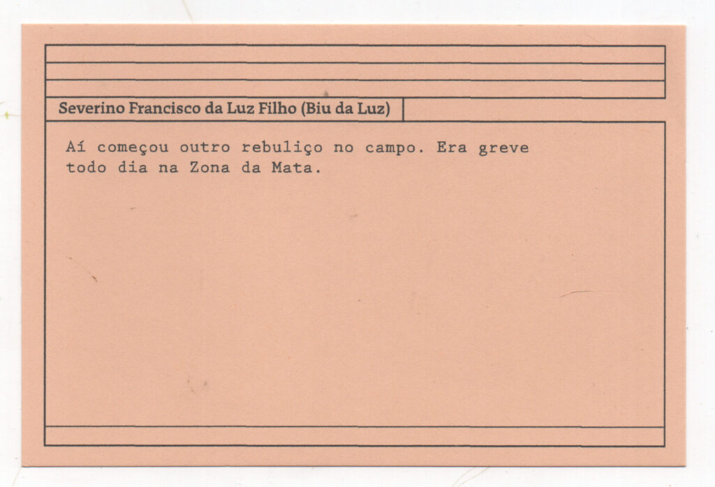 Fotografia de uma ficha horizontal cor de rosa claro com cinco pautas pretas na
parte superior. Abaixo delas, em letras pretas, o depoimento de Severino Francisco
da Luz Filho (Biu da Luz), em letras pretas: “Aí começou outro rebuliço no campo.
Era greve todo dia na Zona da Mata.” No rodapé, duas pautas pretas horizontais.