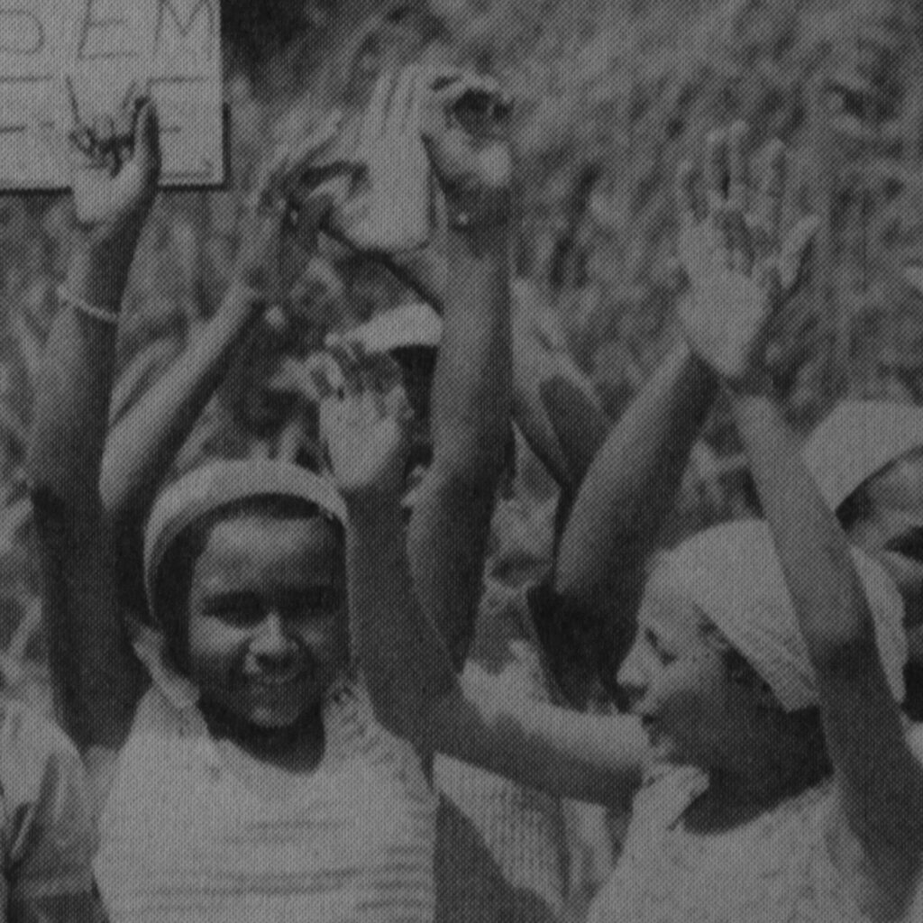 Foto de uma fotografia antiga, preta e branca, na vertical de duas meninas, da
cintura para cima, com os braços levantados para cima. A do lado esquerdo, está de
frente para a foto, sorridente. Ela usa um pano na cabeça, uma pulseira no braço
direito e blusa de alça grossa. De perfil esquerdo, a menina do lado direito, usa
pano na cabeça e blusa de alça grossa. Atrás delas vários braços levantados para
cima.