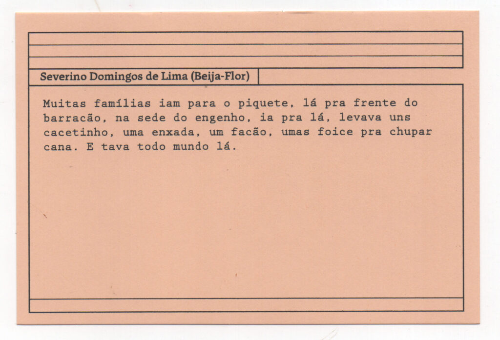 Fotografia de uma ficha horizontal cor de rosa claro com cinco pautas pretas na
parte superior. Abaixo delas, em letras pretas, o depoimento de Severino Domingos
de Lima (Beija-Flor): “Muitas famílias iam para o piquete, lá pra frente do barracão,

na sede do engenho, ia pra lá, levava uns cacetinho, uma enxada, um facão, umas
foice pra chupar cana. E tava todo mundo lá.” No rodapé, duas pautas pretas
horizontais.