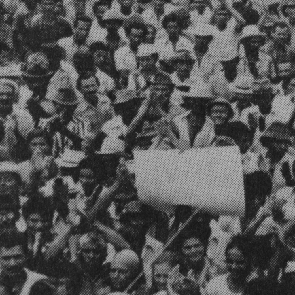Foto de uma fotografia antiga em preto e branco na vertical. Vista aérea de uma
multidão de homens e mulheres em greve. Todos estão voltados para a frente, a
maioria com expressão facial séria. Alguns aplaudem; uma pessoa segura um
cartaz.