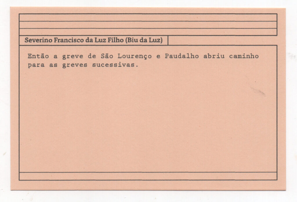 Fotografia de uma ficha horizontal cor de rosa claro com cinco pautas pretas na
parte superior. Abaixo delas, em letras pretas, o depoimento de Severino Francisco
da Luz Filho (Biu da Luz): “Então a greve de São Lourenço e Paudalho abriu
caminho para as greves sucessivas.” No rodapé, duas pautas pretas horizontais.