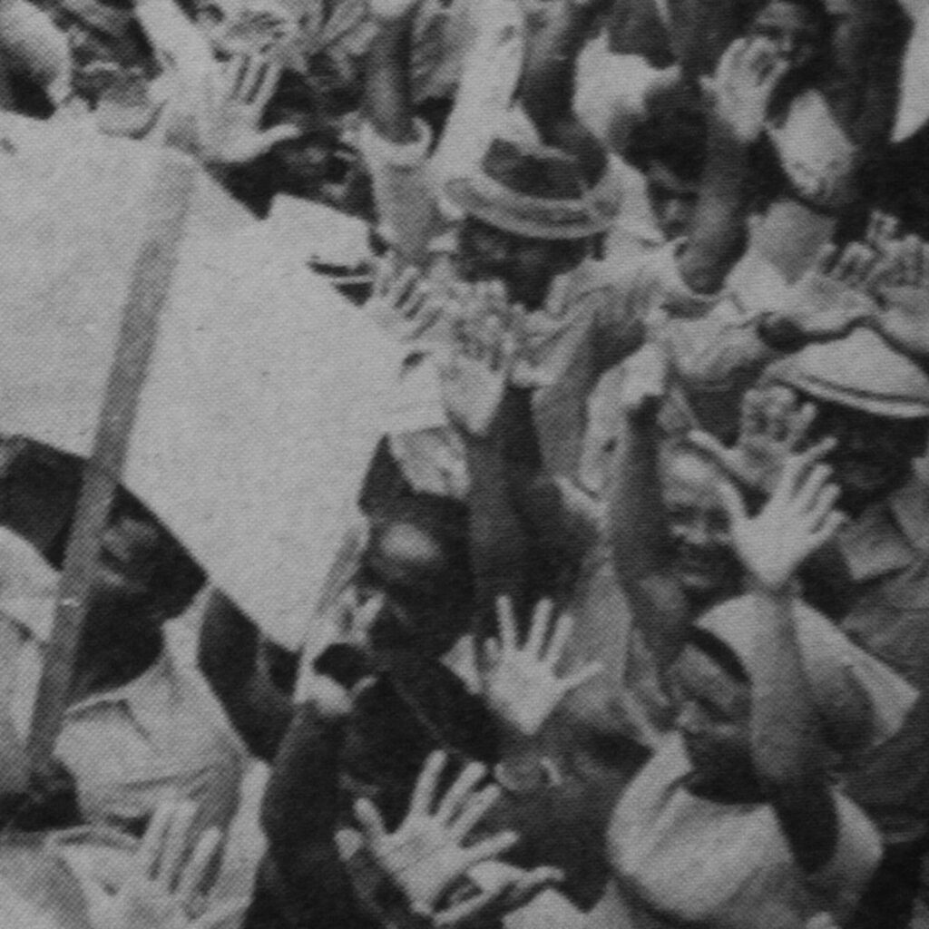 Foto de uma fotografia antiga, preta e branca, na vertical, tirada de cima, de vários
homens e mulheres em pé, com os braços levantados e com as mãos espalmadas.
Do lado esquerdo, um homem levanta com a mão esquerda uma placa branca
fixada em um suporte de madeira.