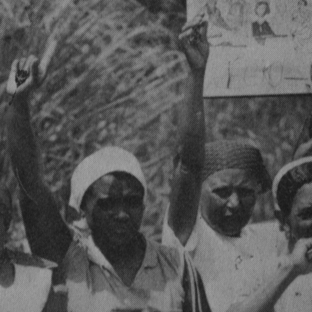Foto de uma fotografia antiga preta e branca na vertical de duas mulheres pretas, da
cintura para cima, lado a lado, de frente para foto, com testas franzidas e lábios
cerrados. A mulher do lado esquerdo, usa pano claro na cabeça, uma blusa de alça
grossa e tem o braço direito levantado, segura um objeto circular e aponta para
cima com o dedo indicador. A do lado direito, usa um pano com detalhes mais
escuros na cabeça e levanta o braço direito com a mão entreaberta. Atrás delas,
galhos e uma placa com desenhos de mulheres e em preto “feto”.