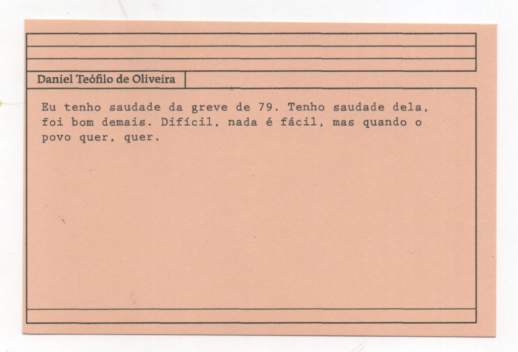 Fotografia de uma ficha horizontal cor de rosa claro com cinco pautas pretas na
parte superior. Abaixo delas, em letras pretas, o depoimento de Daniel Teófilo de
Oliveira: “Eu tenho saudade da greve de 79. Tenho saudade dela, foi bom demais.
Difícil, nada é fácil, mas quando o povo quer, quer.” No rodapé, duas pautas pretas
horizontais.
