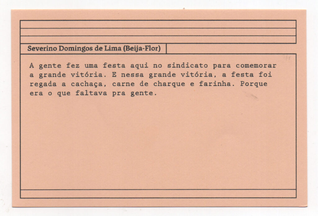 Fotografia de uma ficha horizontal cor de rosa claro com cinco pautas pretas na
parte superior. Abaixo delas, em letras pretas, o depoimento de Severino Domingos
de Lima (Beija-Flor): “A gente fez uma festa aqui no sindicato para comemorar a
grande vitória. E nessa grande vitória, a festa foi regada a cachaça, carne de
charque e farinha. Porque era o que faltava pra gente.” No rodapé, duas pautas
pretas horizontais.