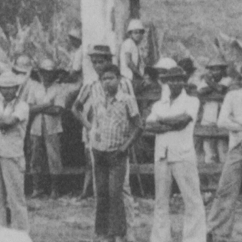 Foto de uma fotografia antiga em preto e branco na vertical. Uma cerca de madeira
divide dois grupos de trabalhadores, sobre a qual há dois homens sentados. Os
trabalhadores à frente da cerca estão de braços cruzados. Entre eles, há um menino
com as mãos nos bolsos da calça que usa.