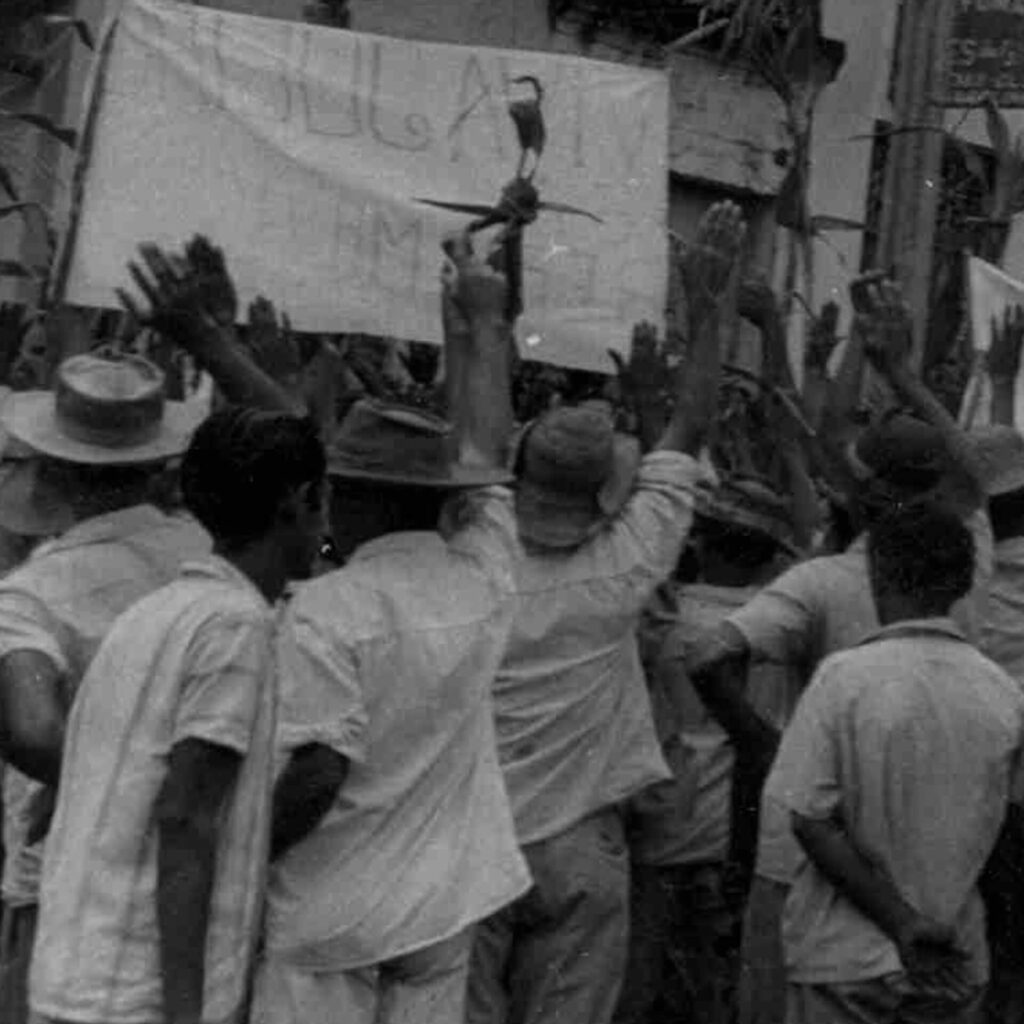 Foto de uma fotografia antiga em preto e branco na vertical. No topo de uma
construção feita com tábuas de madeira, há um cartaz branco com alguns escritos.
À frente do cartaz, trabalhadores estão com os braços erguidos. Um homem segura
no alto um objeto semelhante a uma cruz, com as hastes laterais delgadas e
pontudas.