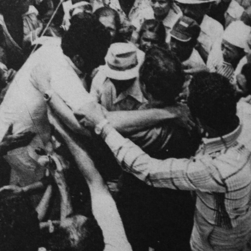 Foto de uma fotografia antiga em preto e branco na vertical. Um grupo faz um cordão de proteção ao redor de um homem de cabelo escuro e camisa clara, que está entre uma multidão de trabalhadores.