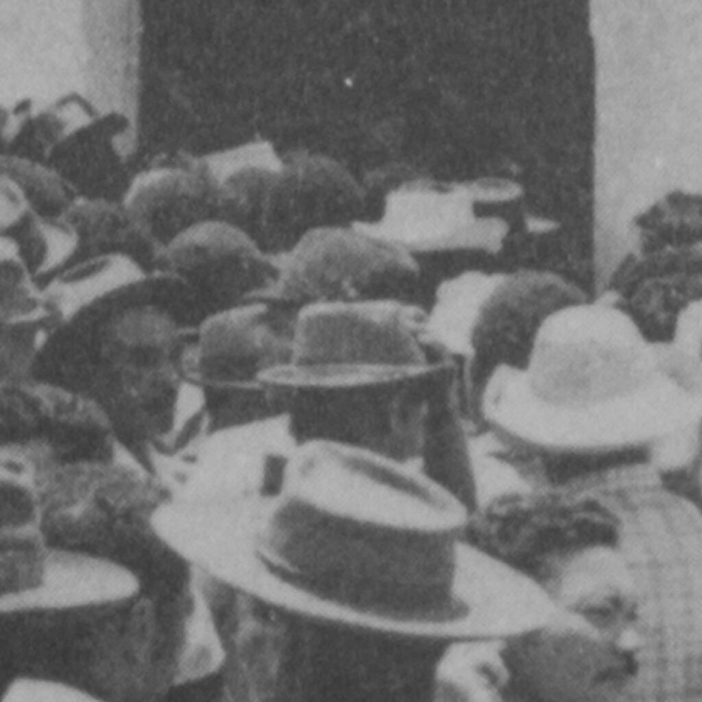 Foto de uma fotografia antiga em preto e branco na vertical com foco no rosto de
trabalhadores, que estão diante da entrada escura de algum lugar de paredes
claras. Eles olham para sentidos opostos, alguns para a entrada escura, outros para
a direita.